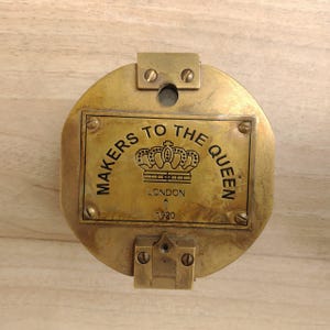 May include: A circular brass plaque featuring a rectangular central plate with the engraved text MAKERS TO THE QUEEN, a crown illustration, LONDON, and the year 1920. The brass surface shows a metallic finish with visible screw fasteners.