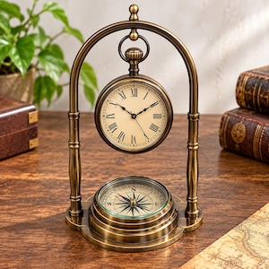 Può includere: Un orologio da scrivania in metallo color ottone sospeso a una cornice ad arco, con un quadrante rotondo con numeri romani e lancette nere. Sotto l'orologio, una bussola circolare è integrata nella base, indicando i punti cardinali.