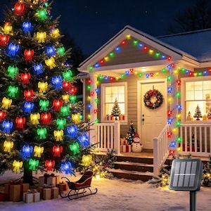 May include: A string of LED lights shaped like small bows in red, green, blue, and yellow, draped over a Christmas tree and the exterior of a house. A black solar panel on a stake is positioned in the foreground to power the decorative lights.