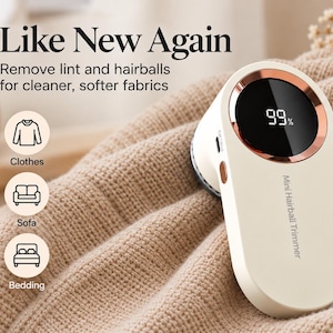 May include: A cream-colored electric fabric shaver with a rose gold circular accent and a digital display showing 99%. The device is labeled Mini Hairball Trimmer and features text stating Like New Again, Remove lint and hairballs for cleaner, softer fabrics, with icons for clothes, sofa, and bedding.