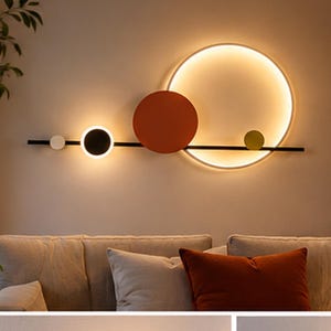 Puede incluir: Un aplique de pared que presenta una barra horizontal negra adornada con cinco elementos circulares de diversos tamaños y colores, incluyendo un gran anillo iluminado, un disco naranja sólido, un disco negro, un pequeño disco blanco y un pequeño disco verde.
