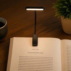 Op de afbeelding: Een zwarte clip-on leeslamp met een flexibele nek en een rechthoekige led-kop die een warm wit licht uitstraalt. De lamp is bevestigd aan de bovenkant van een open boek en verlicht de pagina met de tekst Part Two An Unexpected Turn.