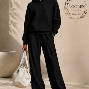 May include: A black hooded sweatshirt with a front kangaroo pocket and matching black wide-leg trousers with a drawstring waist. A cream-colored leather handbag is held at the side. The text NOOREN STYLES FASHION FOR EVERY YOU EST 2024 is visible.