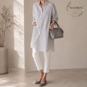 Pode incluir: Uma camisa branca de manga comprida com riscas verticais pretas, gola e fecho de botões. A camisa é combinada com calças brancas lisas e sabrinas cinzentas. Uma mala cinzenta é segurada na mão. O texto diz NOOREN STYLES.