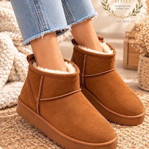May include: A pair of ankle-height brown suede boots with a thick, flat rubber sole and a plush white faux-fur lining. The boots feature a pull tab at the heel and visible stitching along the seams. The text NOOREN STYLES FASHION FOR EVERY YOU EST. 2024 is visible in the upper right corner.