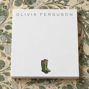 May include: A white rectangular notepad featuring the name OLIVIA FERGUSON printed in dark green capital letters at the top and a small illustration of a pair of dark green rubber rain boots positioned at the bottom center of the page.