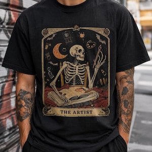 Pode incluir: Uma t-shirt preta de manga curta com uma estampa gráfica de um esqueleto sentado a uma secretária com um livro aberto, um pincel e material de arte. O design é emoldurado como uma carta de tarot com o texto THE ARTIST na parte inferior.