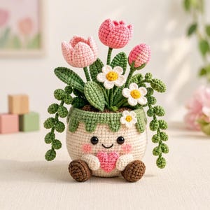 Könnte beinhalten: Eine gehäkelte Topfpflanze mit einem beigen, lächelnden Topf, rosa Wangen und einem kleinen rosa Herz. Die Pflanze besteht aus rosa Tulpen, weißen Gänseblümchen mit gelber Mitte und grünen Ranken mit kleinen runden Blättern aus Garn.