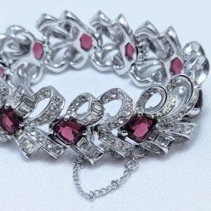 May include: A silver-toned link bracelet featuring repeating bow-shaped motifs encrusted with clear baguette and round rhinestones. Each link is centred with a square-cut deep magenta gemstone. The bracelet includes a safety chain.