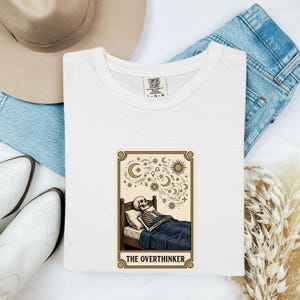 May include: A white t-shirt featuring a graphic of a skeleton lying in bed under a starry sky with celestial symbols. The text "THE OVERTHINKER" is printed below the illustration. The shirt has a Comfort Colors label at the inner neckline.