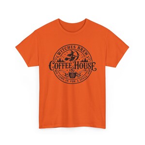 May include: A bright orange short-sleeved t-shirt featuring a black circular graphic on the chest. The design includes a witch on a broomstick, a steaming coffee cup with a pentagram, and the text Witches Brew Coffee House Come In For A Spell.