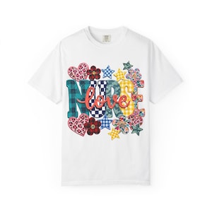 May include: A white short-sleeve t-shirt featuring a graphic design with the word NURSE in large, multi-patterned block letters, overlaid with the word love in red script. The design includes colourful hearts, stars, and flowers in various prints.