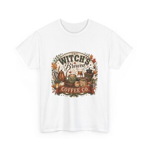 May include: A white short-sleeve t-shirt featuring a central graphic with the text "WITCH'S BREWED COFFEE CO." The design includes a copper kettle, a green mug, a glass carafe, a coffee grinder, and autumn leaves in shades of orange and brown.