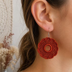 May include: A round, rust-colored crochet earring featuring a central circular opening and a scalloped, petal-like outer edge. The earring is attached to a silver-toned metal hook, showcasing intricate thread work and a vibrant, warm-toned finish.