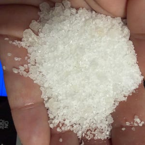 May include: A pile of coarse, translucent white crystalline granules resting in a palm. The crystals vary in size and shape, reflecting light with a bright, clear appearance. The texture is granular and irregular, consistent with sea salt or mineral crystals.