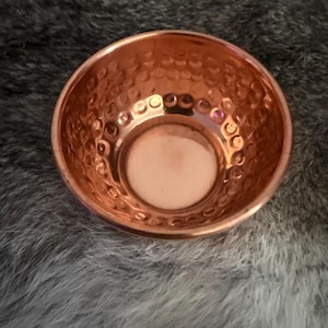 May include: A small, round, metallic bowl with a polished copper finish. The interior walls of the bowl feature a distinct hammered texture, creating a series of small, circular indentations that catch the light, while the base remains smooth.