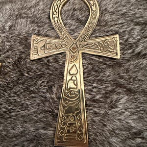 May include: A metallic gold-toned ankh symbol featuring intricate engraved hieroglyphic-style carvings across its entire surface. The cross-shaped object has a rounded loop at the top and a long vertical base, with detailed etchings of various symbols.