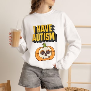 May include: A white long-sleeve sweatshirt featuring the yellow text I HAVE AUTISM in a bold block font with a black drop shadow. Below the text is a graphic of an orange pumpkin with a white skull face and a green stem on the top centre.
