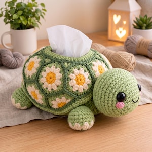 May include: A crochet turtle tissue box cover featuring a green shell constructed from hexagonal motifs with white and yellow daisy patterns. The turtle has a light green head with large black eyes, pink cheeks, and a stitched smile, holding a tissue.