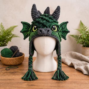 Peut inclure: Un bonnet dragon au crochet dans des tons de vert forêt et foncé, avec deux cornes gris foncé, des détails en écailles texturées et des ailes latérales. Le bonnet comprend un museau, des yeux jaunes et noirs, et deux longues tresses terminées par des pompons.