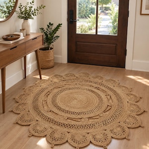 May include: A round, tan-colored woven jute rug featuring a central circular pattern surrounded by a decorative scalloped border with intricate braided details. The rug is placed on a light-colored wooden floor in an entryway setting.