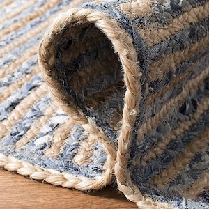 May include: A braided area rug featuring alternating rows of natural tan jute fibers and strips of blue denim fabric. The rug is partially folded over to reveal the thick, woven texture of the braided construction and the underside of the material.