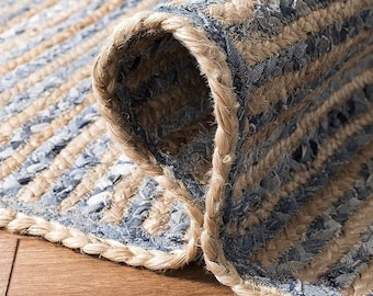 May include: A braided area rug featuring alternating rows of natural tan jute fibers and strips of blue denim fabric. The rug is partially folded over to reveal the thick, woven texture of the braided construction and the underside of the material.