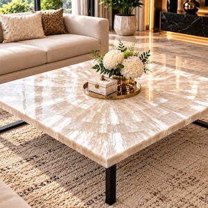 May include: A square coffee table featuring a polished, light-colored stone surface with a radial, sunburst-like pattern of inlaid segments. The table is supported by four black metal legs and holds a small decorative box and flowers.