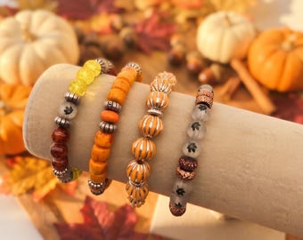 May include: Four beaded stretch bracelets displayed on a cylindrical stand. The bracelets feature a variety of beads including yellow crackle glass, solid orange, ribbed orange with silver accents, and frosted glass beads with black maple leaf prints.