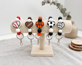 May include: Five silicone bead keychains featuring heart-shaped charms with sports themes including baseball, football, basketball, soccer, and volleyball. Each keychain has a metal split ring and a lobster clasp attached to a string of beads.