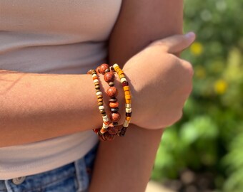 May include: A multi-strand bracelet featuring a variety of beads in shades of brown, orange, and cream. The design includes round wooden beads with visible grain, smaller dark brown spherical beads, and square-shaped beads in yellow and cream tones.