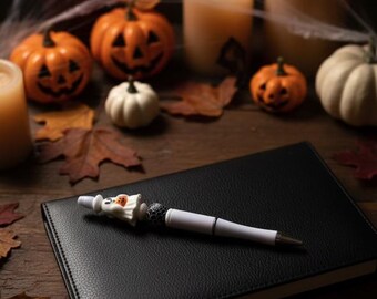 May include: A white ballpoint pen featuring a small ghost figurine holding a pumpkin, a black bead with a white spiderweb pattern, and a silver-toned tip, resting on a black rectangular notebook with a textured cover.