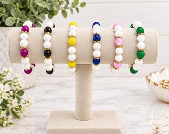 May include: Six beaded bracelets displayed on a beige T-bar stand. Each bracelet features white beads alternating with coloured beads in magenta, black, yellow, blue, pink, and green, all separated by small gold-toned metal spacers between each bead.