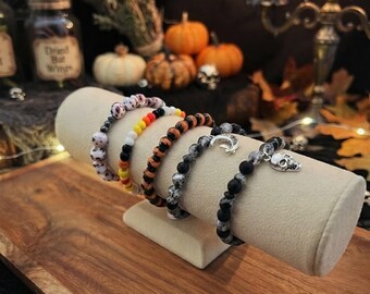 May include: A set of five beaded bracelets displayed on a beige cylindrical stand. The bracelets feature various designs including white with black spots, candy corn colours, orange and black stripes, grey speckled beads with a moon charm, and black beads with a silver skull charm.