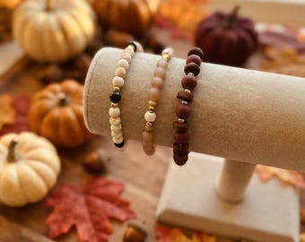 May include: Three beaded bracelets displayed on a beige T-bar stand. The first bracelet features white and tan marbled beads with a single black bead. The second has light tan frosted beads. The third consists of dark maroon matte beads.