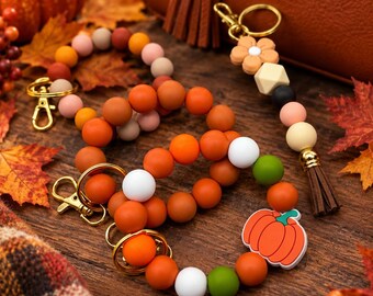 May include: Three circular silicone bead keychains featuring a mix of orange, white, and green beads, with one keychain including a pumpkin-shaped charm. A separate linear keychain features a flower charm, geometric bead, and a brown tassel.