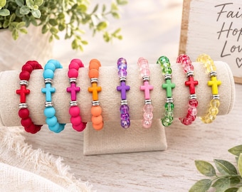May include: A collection of nine stretch bracelets displayed on a beige stand. Each bracelet features round beads in various colours including red, blue, pink, orange, purple, green, and yellow, accented by a central cross charm and silver spacers.