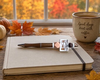 May include: A brown wooden pen featuring a square bead with the text "BEAUTY COMES IN ALL SHAPES & SIZES" alongside illustrations of coffee drinks, accented by a peach-coloured geometric bead and smaller dark brown and white spherical beads.