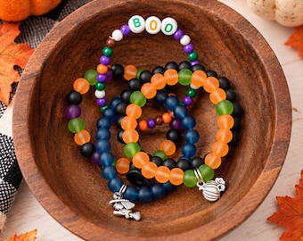 May include: A collection of beaded bracelets featuring matte and translucent beads in orange, green, purple, black, and blue. One bracelet includes white letter beads spelling out BOO. Two bracelets feature silver-toned charms of a witch and a pumpkin.