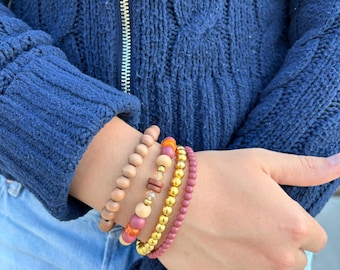 May include: A stack of four beaded bracelets featuring a variety of round beads in shades of matte mauve, polished gold, and natural wood, along with accent beads in orange and clear crystal, worn together on a wrist.