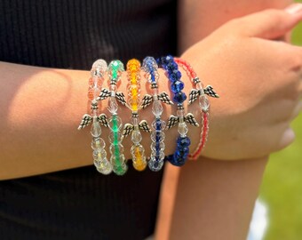 May include: A set of six elastic beaded bracelets featuring faceted glass beads in clear, green, orange, light blue, dark blue, and red. Each bracelet includes a central silver-toned metal charm shaped like a pair of angel wings.