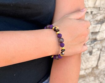 May include: A beaded bracelet featuring alternating purple textured beads, brown mottled round beads, and small gold-toned metallic spacers. The bracelet includes a small gold-toned crescent moon charm integrated into the bead sequence.