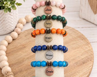 May include: A vertical display of seven beaded stretch bracelets, each featuring a round metal charm engraved with words including Hope, Love, Faith, Dream, Blessed, Create, and Peace. The bracelets are made of various coloured stone beads.