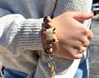 May include: A wristlet keychain featuring a series of round, matte-finish beads in shades of brown and tan. The keychain includes a silicone charm shaped like a Highland cow and a metal split ring with a small, light brown faux leather tassel.