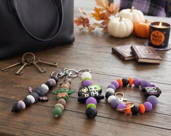 May include: Three silicone bead keychains and one silicone bead wristlet keychain. The keychains feature a bat, a green alien character, and two figures with black and green hair. The wristlet includes a charm with the text Hocus Pocus.