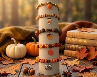 May include: A stack of five beaded bracelets wrapped around a cylindrical display. The bracelets feature charms including an orange pumpkin, a coffee cup, an acorn, an orange maple leaf, and a silver-toned jack-o'-lantern pumpkin charm.