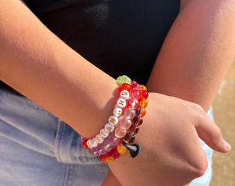 May include: A stack of three beaded bracelets featuring a variety of colourful translucent and opaque beads. One bracelet includes white letter beads spelling out the word "STAHHS" in black text, while another features a small black charm.