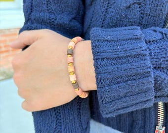 May include: A beaded bracelet featuring alternating flat disc-shaped beads in shades of peach, light pink, and dark brown, interspersed with small, round, metallic gold-toned spacer beads arranged in a repeating pattern around the wrist.