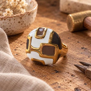 May include: A gold ring featuring a rounded, chunky face with an inlaid geometric pattern of smooth white and dark gray enamel sections. The metal surface is polished and reflective, creating a contrast between the gold and the enamel.