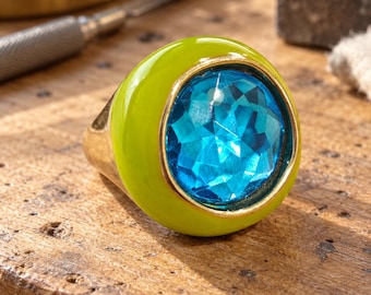 Può includere: Un anello color oro caratterizzato da un'ampia fascia liscia in smalto verde lime e da una grande pietra preziosa rotonda sfaccettata di colore blu brillante, incastonata in una montatura color oro su una superficie di legno ruvida.